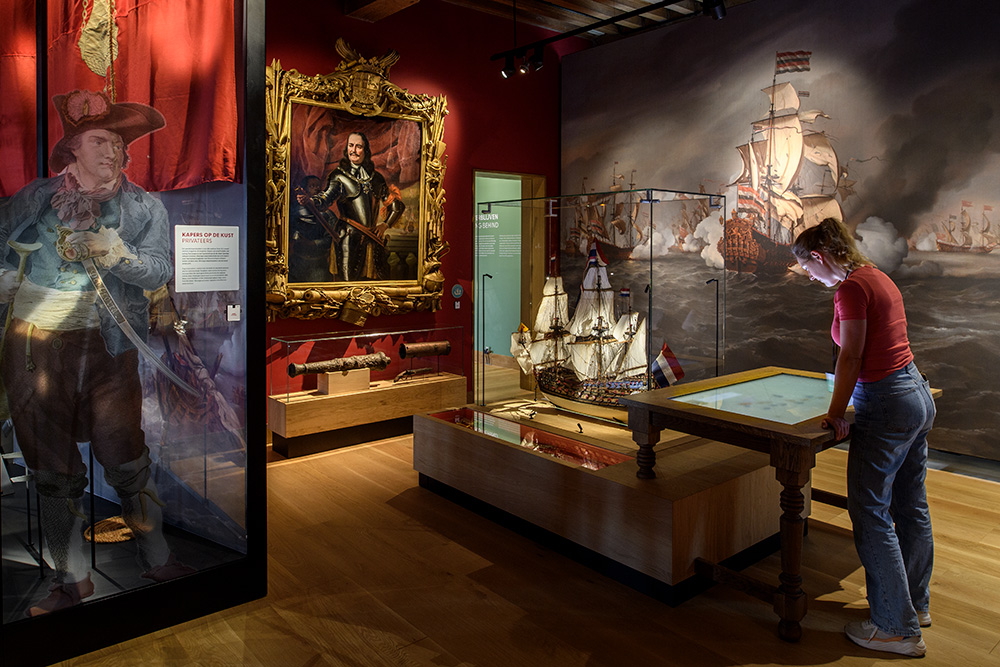Een vrouw kijkt naar een installatie in een museumruimte. De ruimte bevat onder andere een groot schilderij van een 17e-eeuws schip. Een in een gouden lijst ingelijst schilderij van Michiel de Ruyter. Een schaalmodel van een schip en een klein, oud kanon.
