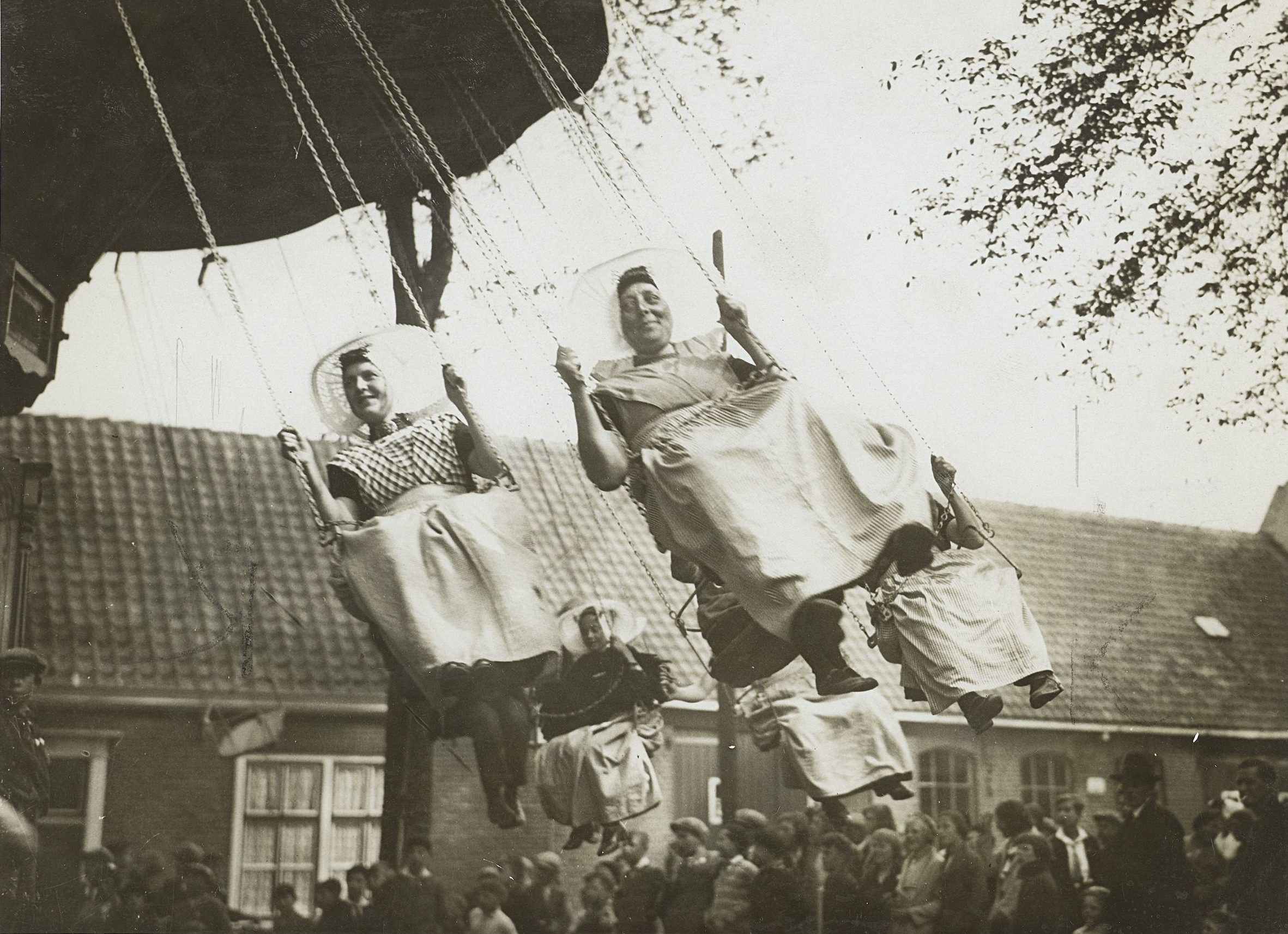 Een zwartwit foto van een zweefmolen die in beweging is. In de zweefmolen zitten vijf vrouwen in klederdracht. Op de achtergrond staat een groep mensen toe te kijken.