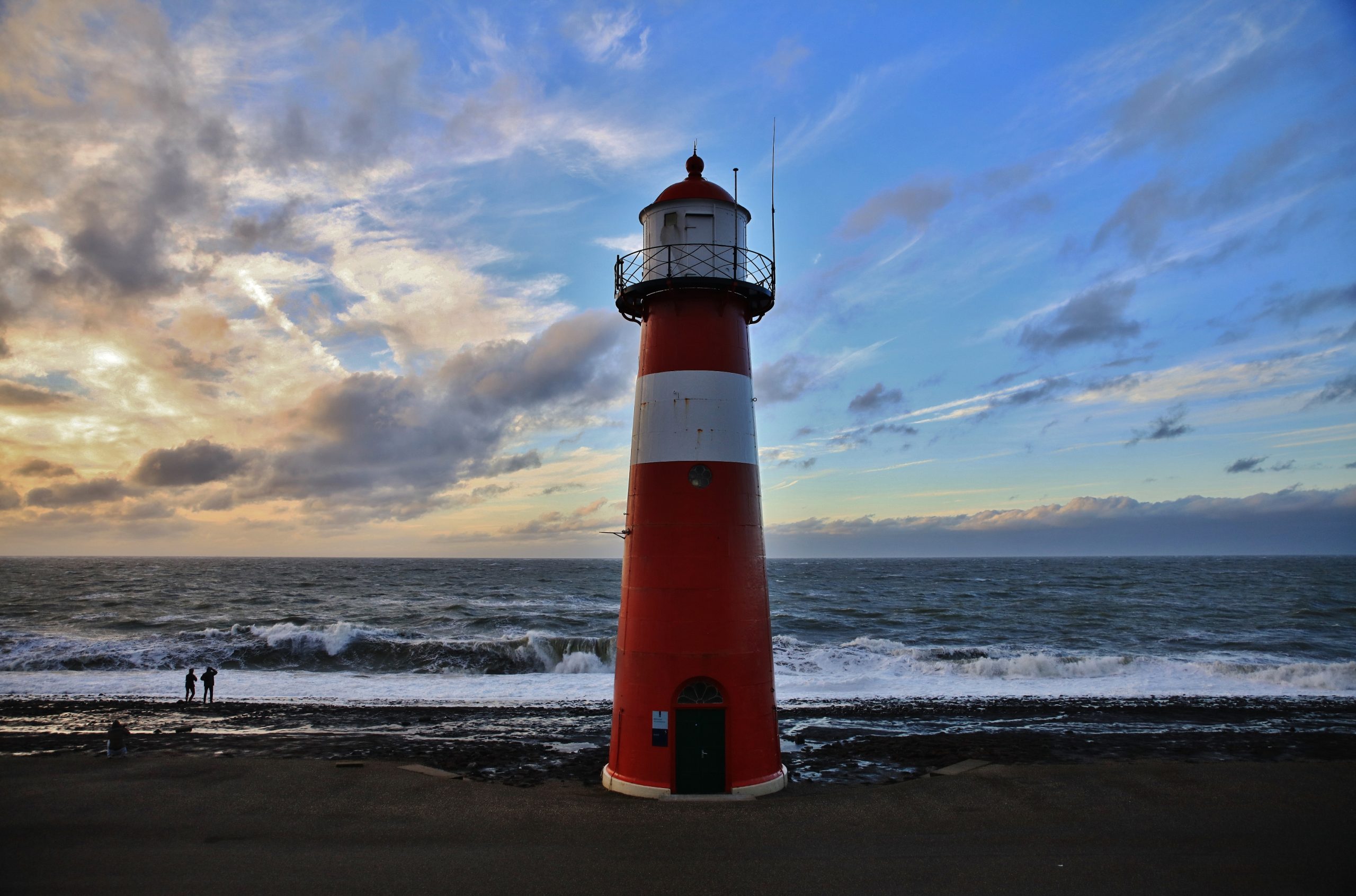 Een gietijzeren rood-witte vuurtoren met op de achtergrond een woeste zee met daarboven een typisch Nederlandse lucht met blauw en wolken. De zon is niet te zien en lijkt aan het licht te zien onder te gaan. Aan de rand van het water staan twee mensen.