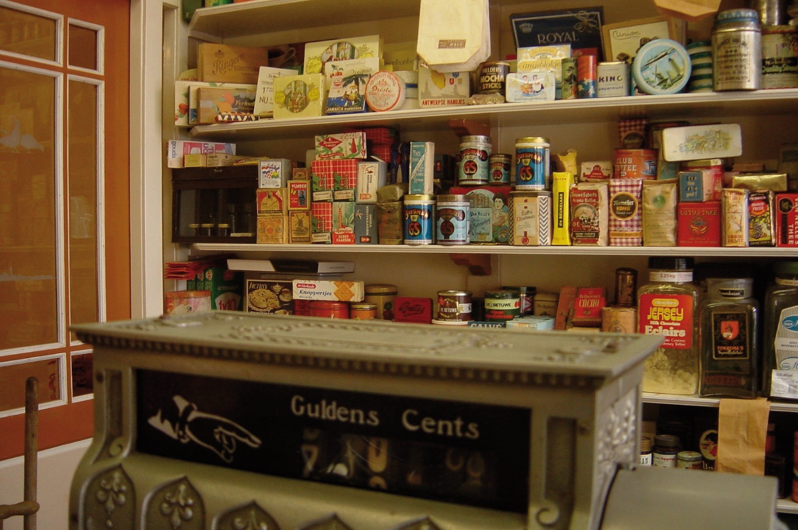 De kassa en voorraad van een ouderwetse supermarkt. De kassa op de voorgrond is van een versierd metaal. Een vinger wijst naar cijfers onder de woorden Guldens en Cents waar kan worden aangegeven hoeveel de klant moet afrekenen. Het assortiment achter de toonbank bestaat uit koffie, thee, suiker, koekjes en ander voedsel: samen vormen ze een rijkgekleurd geheel van blikken, zakken en dozen.