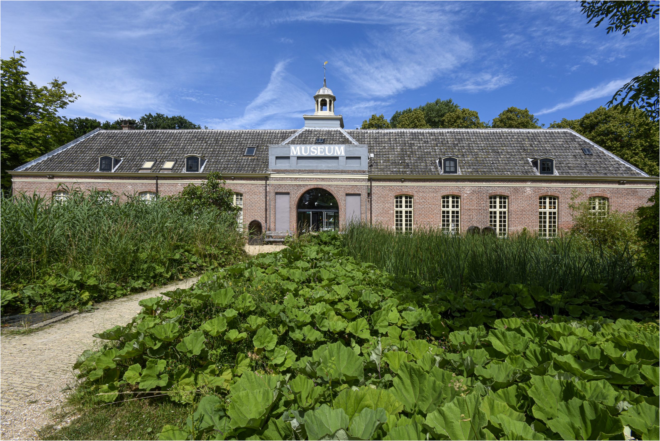 Vooraanzicht van een langwerpig gebouw met een traditionele bakstenen gevel en een symmetrisch zadeldak. Op de voorgrond veel groen en een pad dat naar de ingang van het gebouw leidt. Boven de glazen ingang van het gebouw is op de daklijst het woord MUSEUM te lezen. boven de daklijst is een torentje met een rondvormige koepeltje te zien.