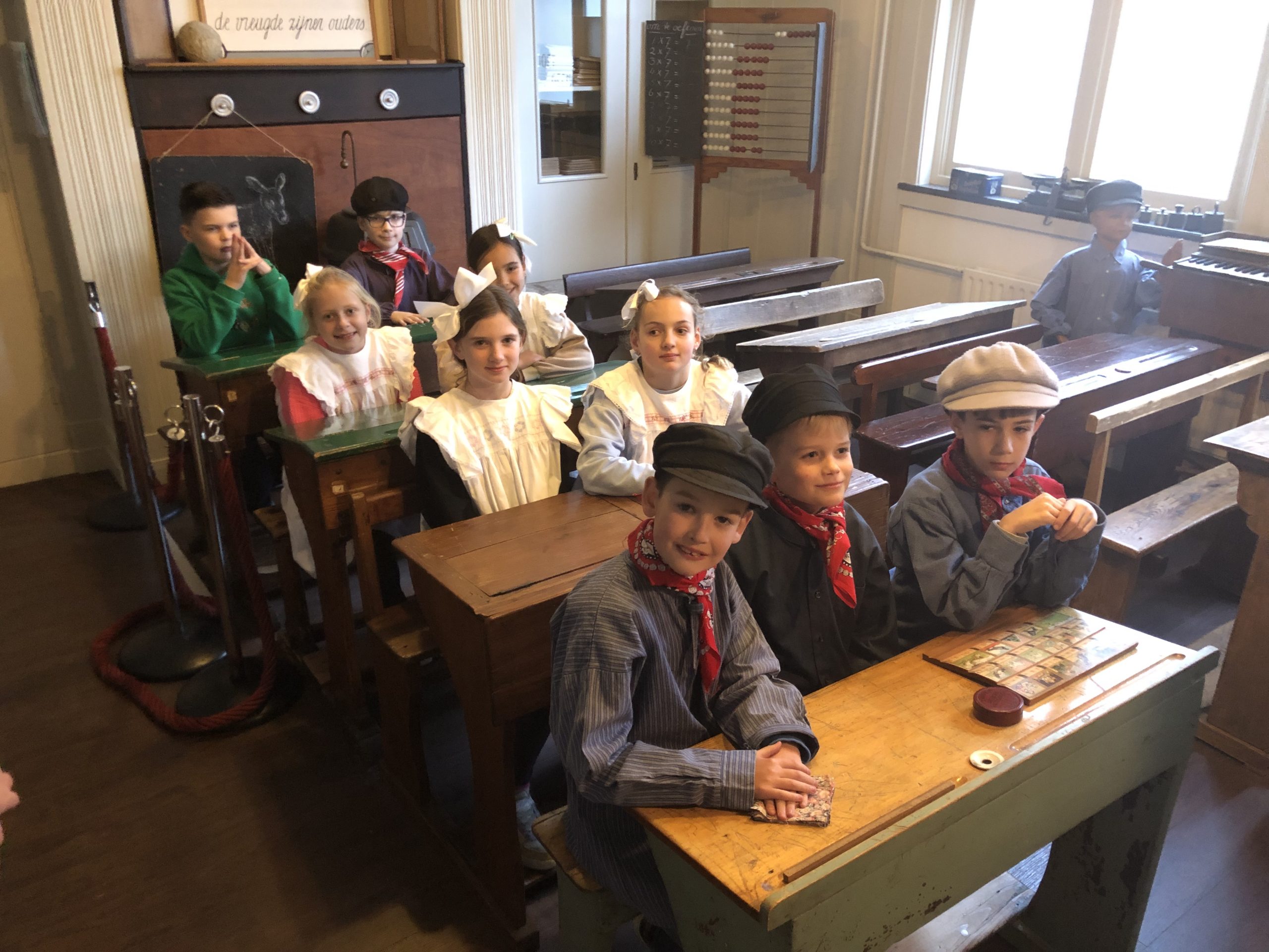 Oud klaslokaal met schoolbankjes. in Museum Schooltijd Terneuzen. Negen kinderen in historische kleding bezig met een schrijfopdracht op een leitje. Op de achtergrond is onder andere een telraam en loden gewichtjes te zien.