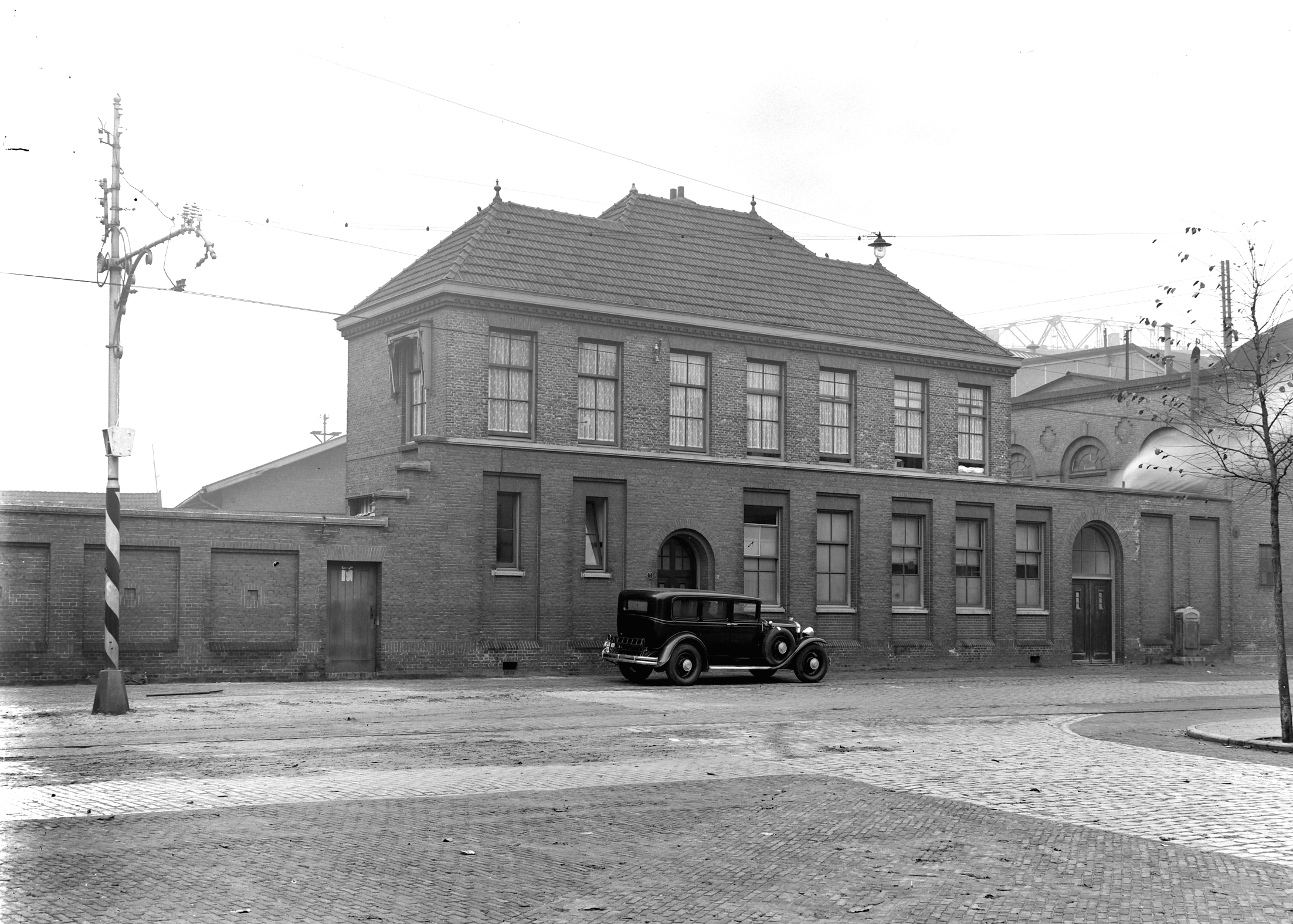 Zwart-witfoto van een statig bakstenen gebouw van twee verdiepingen met zadeldak en symmetrische raampartijen. Voor het gebouw staat een klassieke personenauto uit de jaren 1930 geparkeerd op een geplaveide straat. Rechts op de achtergrond zijn industriële gebouwen zichtbaar. Een kale boom aan de rechterkant duidt op een winter- of herfstseizoen.