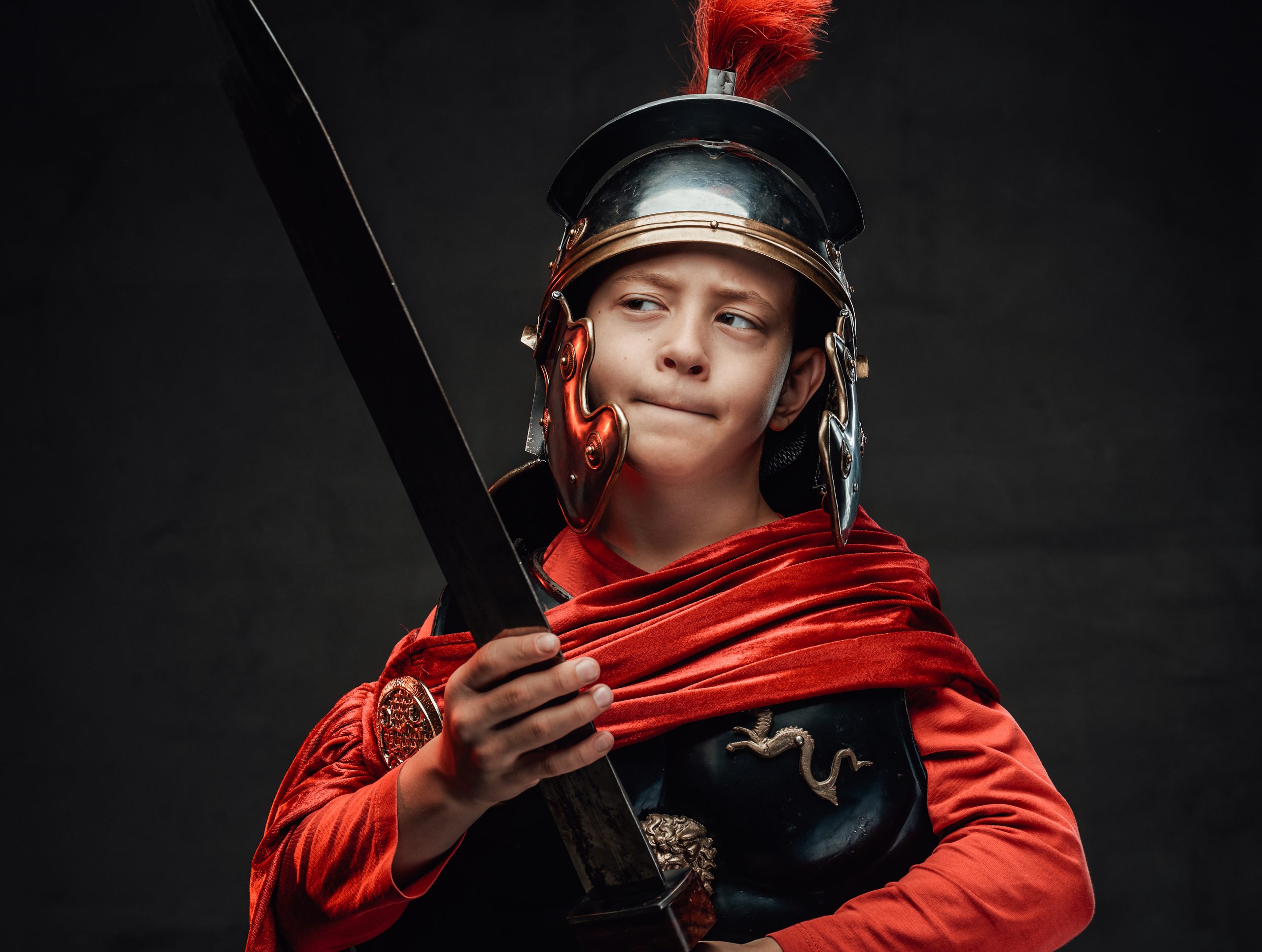 Gekleed in een donker harnas en een rode cape poseert de kleine krijger met zijn zwaard in zijn hand en kijkt ernaar op een donkere achtergrond.