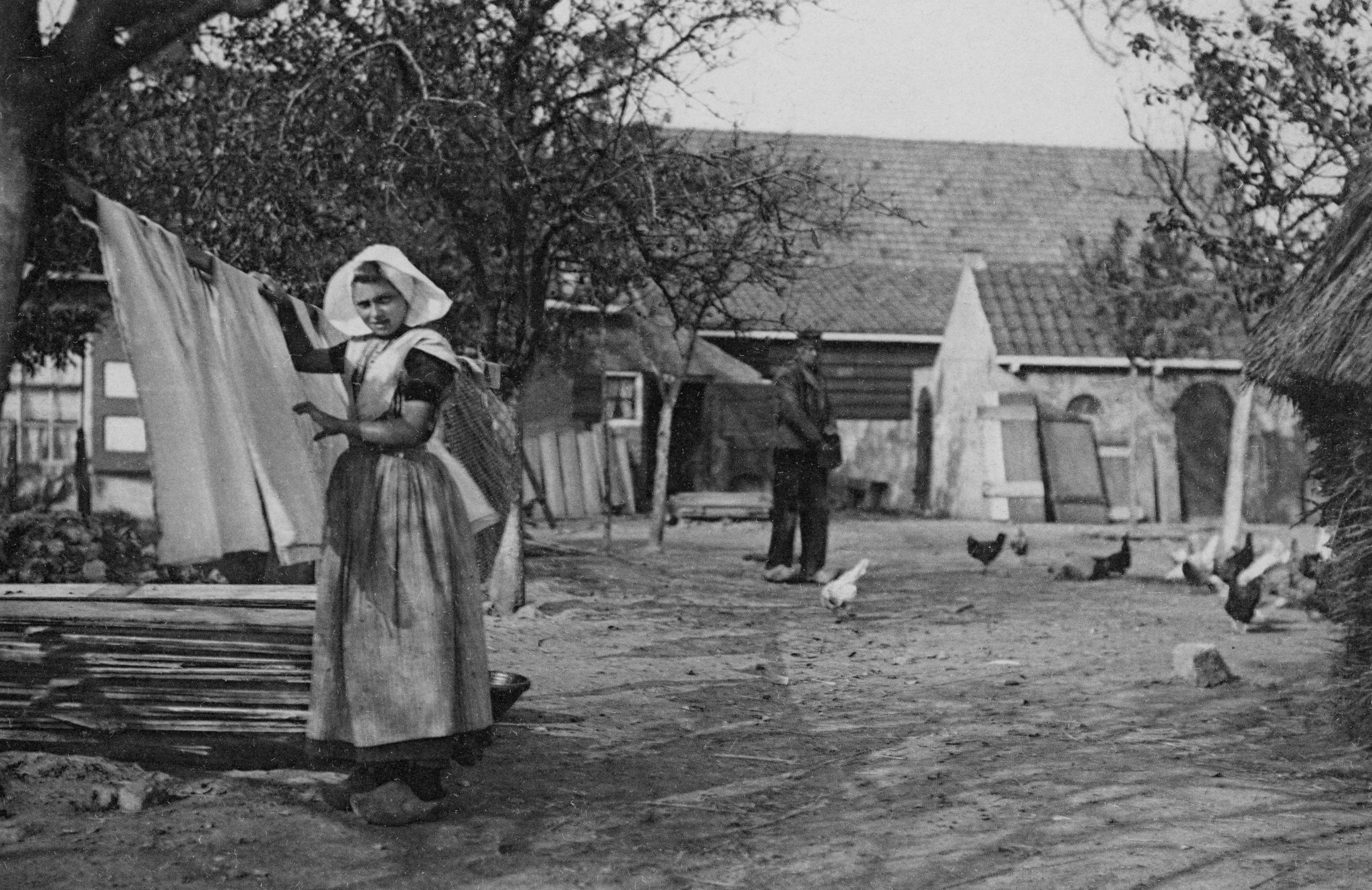 Een vrouw in klederdracht hangt de was op en een man voert kippen, op het erf van een boerderij aan de Nieuwlandseweg in Nieuw- en Sint Joosland. Ca. 1920.