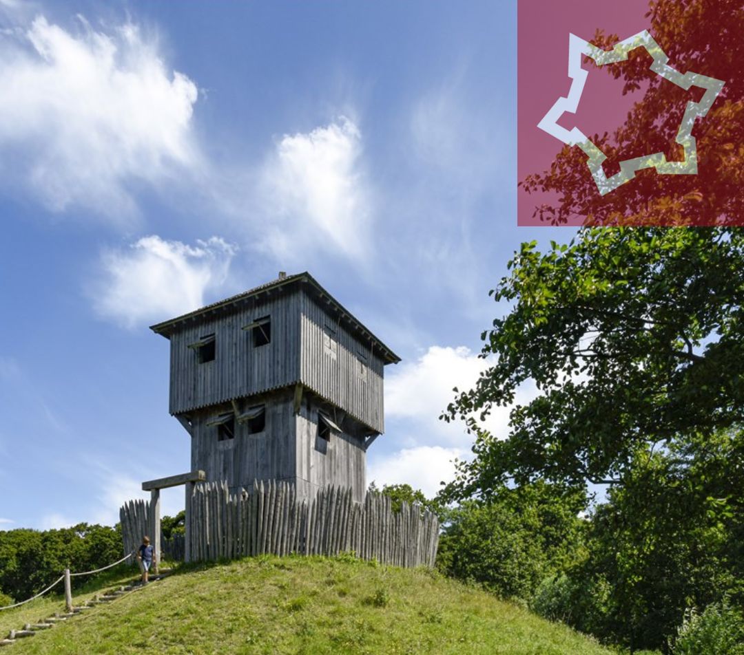 Foto van een mottekasteel. Dit is een houten gebouw van twee verdiepingen omringd door houten palen op een groene heuvel. Recht bovenin het icoon van onze Erfgoedlijn Sterk Zeeland. Dit is een rode tegel met in het wit een ronde stadswal met zes driehoekige uitstulpingen.