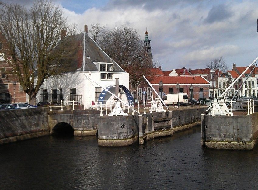 Een historische watermolen en sluis met witte bruggen in een kanaal in Middelburg, Nederland. Op de achtergrond zijn traditionele bakstenen gebouwen met rode daken en een toren met een spits zichtbaar onder een bewolkte lucht.