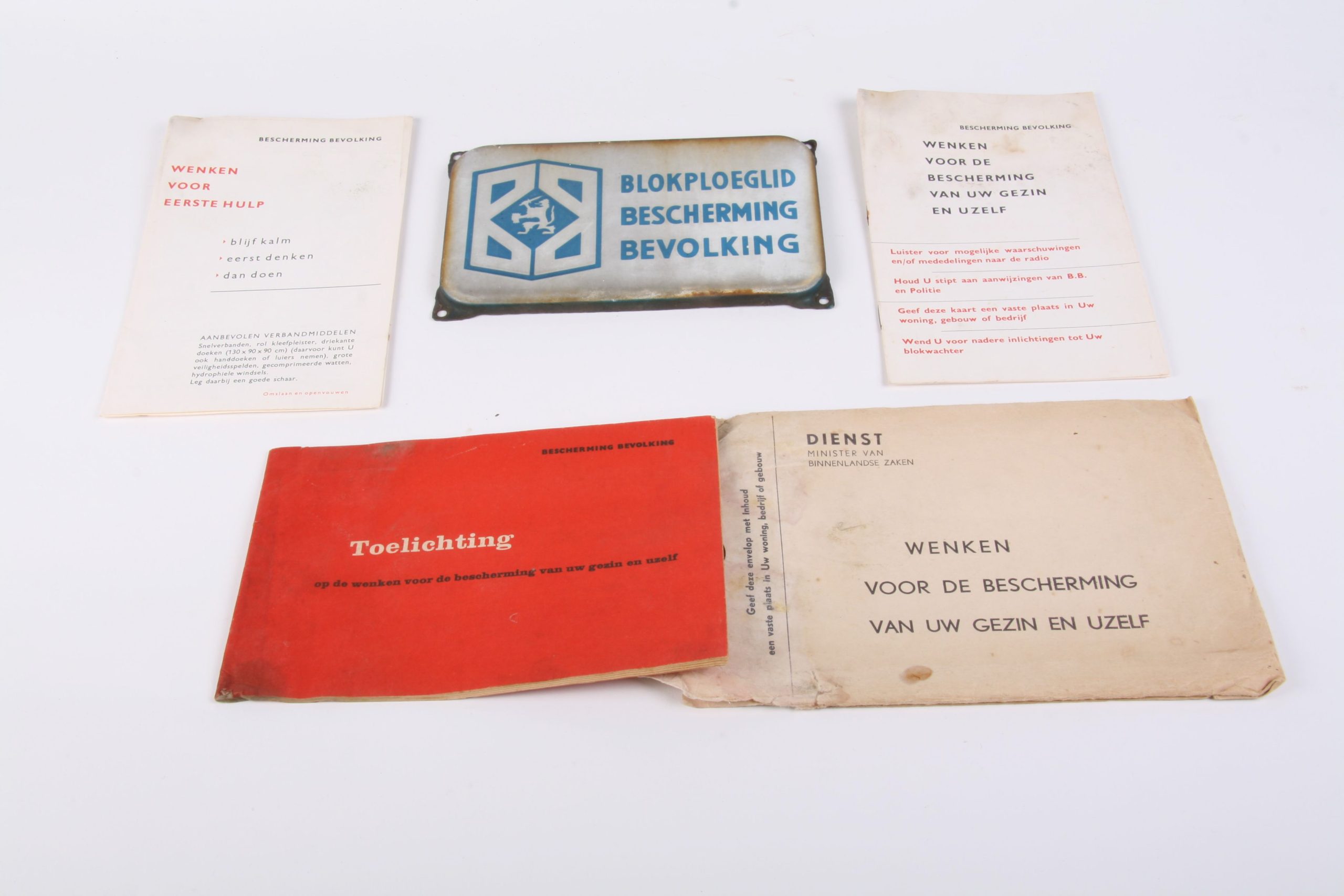A: geëmailleerd naambord "Blokploeglid Bescherming Bevolking" B: enveloppe met zwarte opdruk van de dienst minister van Binnenlandse Zaken: wenken voor de bescherming van uw gezin en uzelf, inhoudende: C: informatieboekje 'wenken voor eerste hulp' D: informatieboekje: wenken voor de bescherming van uw gezin en uzelf E: informatieboekje: toelichting op de wenken van de bescherming van uw gezin en uzelf