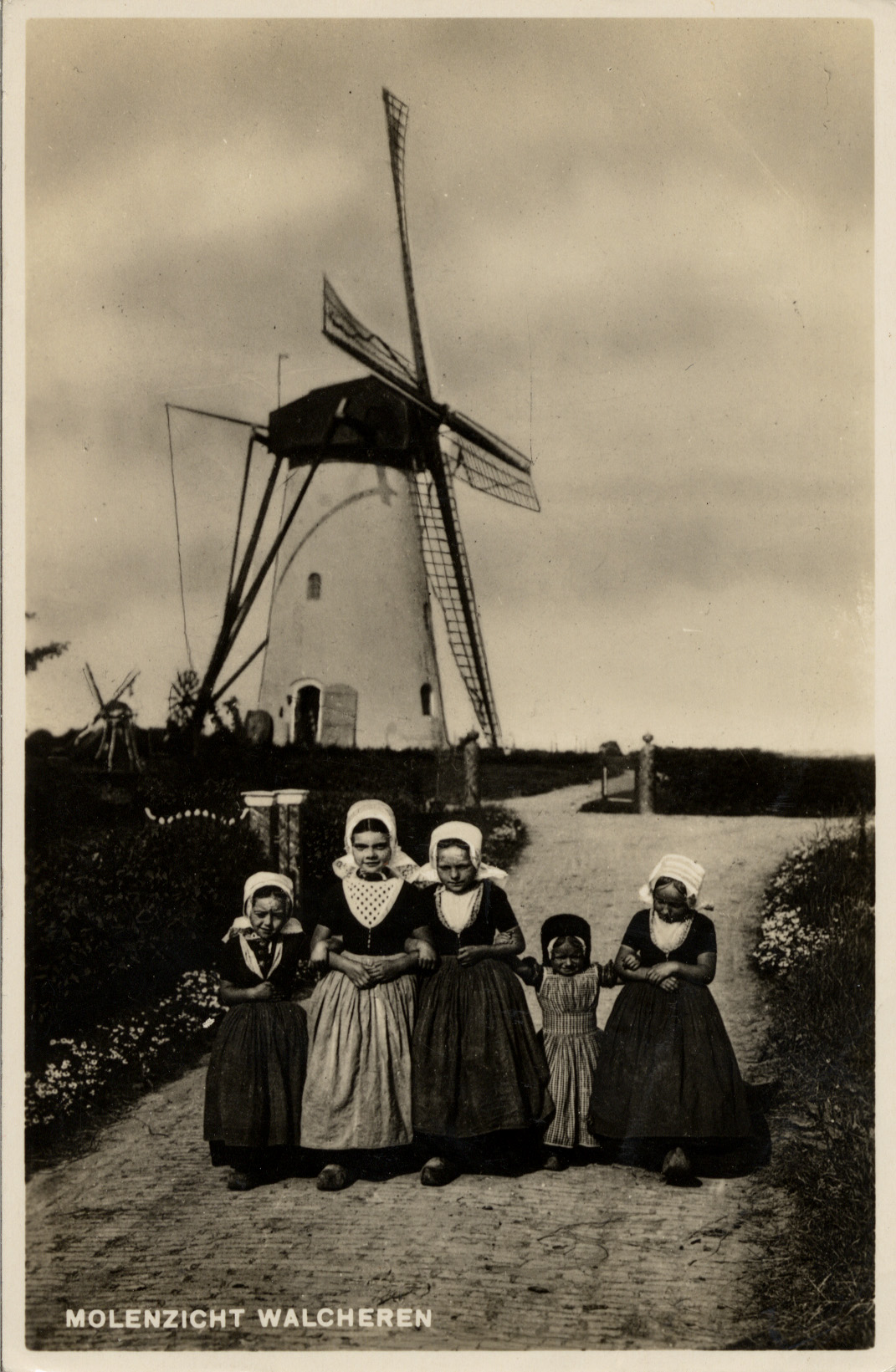 Zwart-witte prentbriefkaart met op de voorgrond vijf meisjes in klederdracht en op de achtergrond een molen.