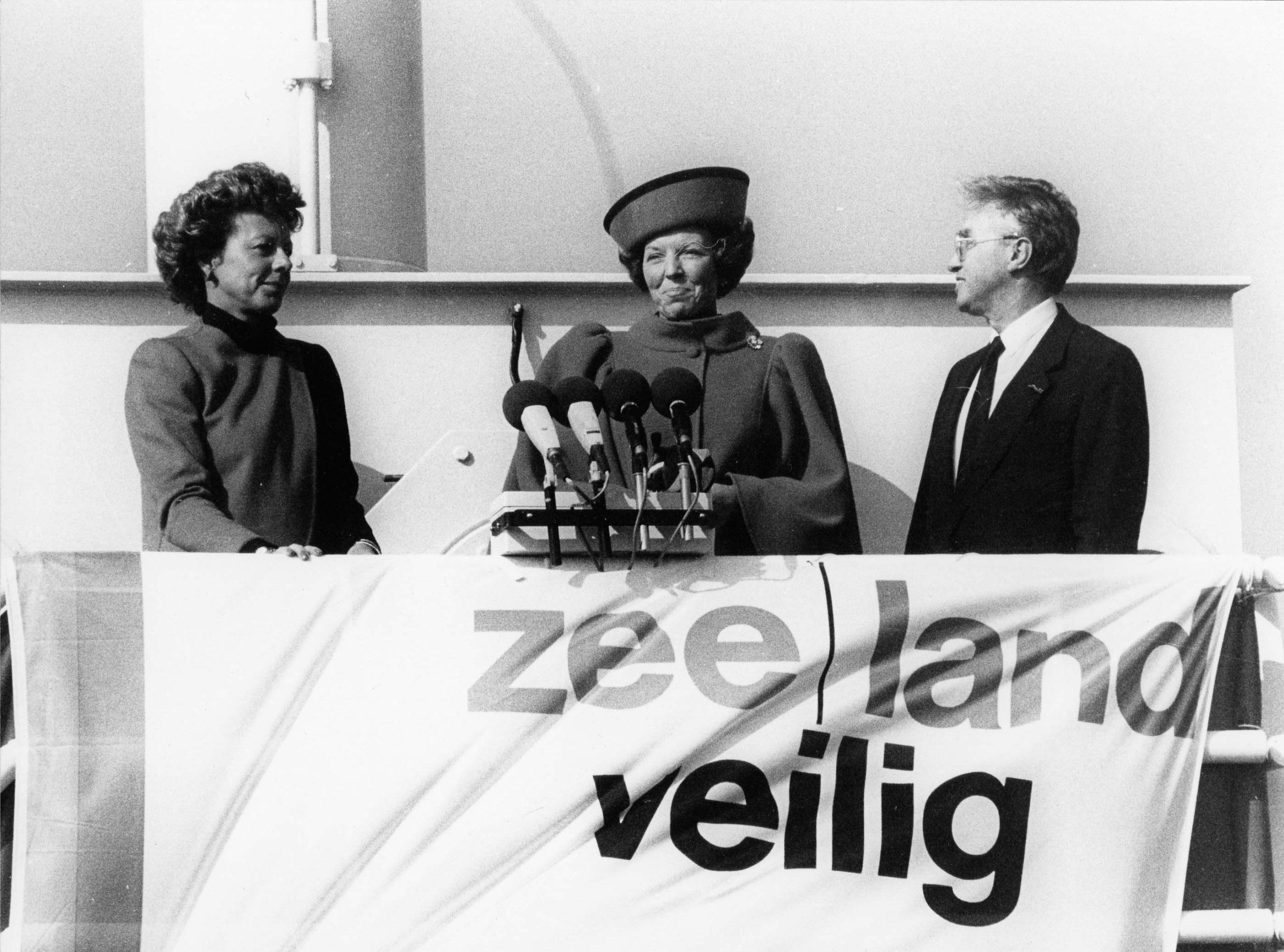 Een zwart-witte foto van een man en een vrouw die kijken naar Koningin Beatrix die tussen hen in staat. Voor de koningin, gekleed in een jas met pofschouders en een toque-achtige hoed, staan vier microfoons. Ook hangt er een vlag met daarop de tekst zee|land veilig.