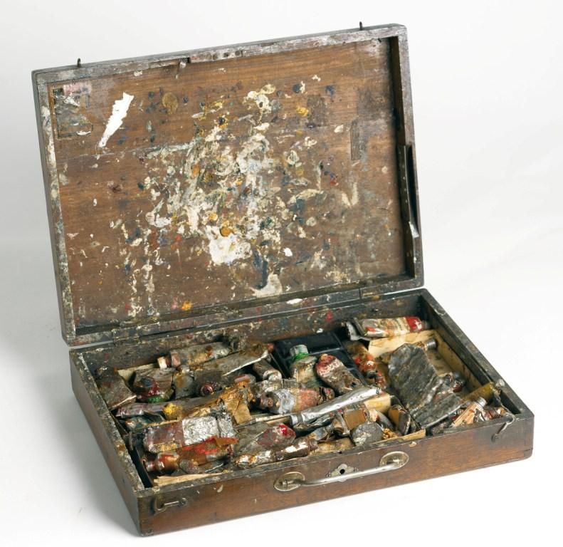 Een foto van een houten schilderkist op een witte achtergrond. De kist staat open en is erg rommelig. Je kunt wat tubes onderscheiden, maar verder vrij weinig.