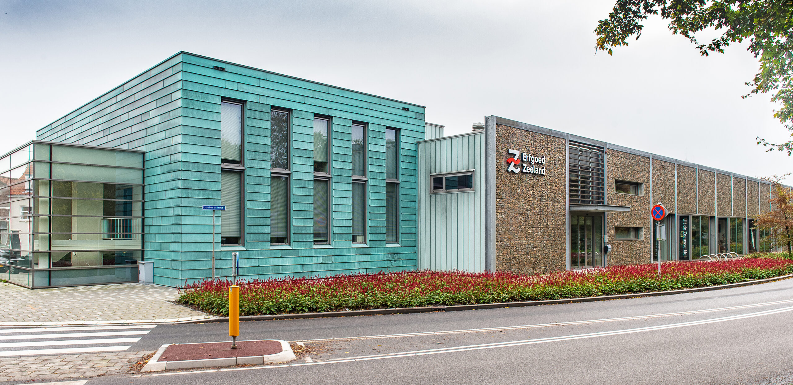 Het kantoor van Erfgoed Zeeland aan de kant van de weg. Links een glazen uitbouw, midden een met blauwkleurige leien vierkant gebouw en rechts een langwerpig gebouw met een stenen uiterlijk.