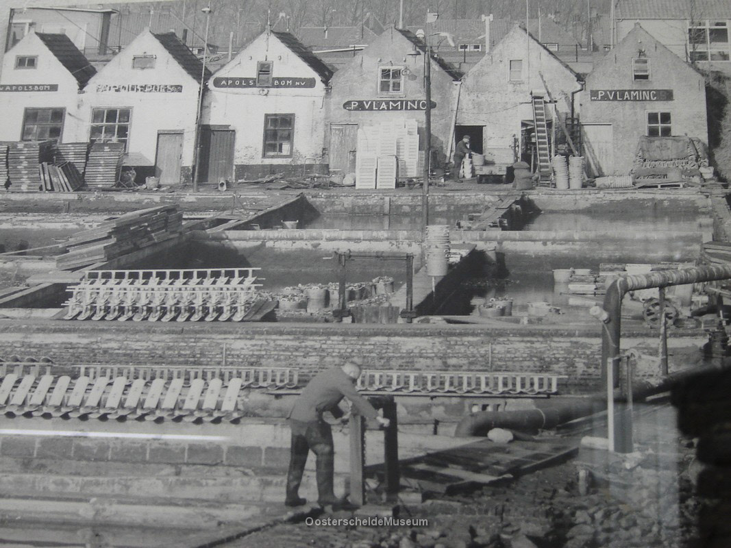 Zwart-wit foto van de oesterputten en inpakloodsen in Yerseke. Op de voorgrond is een man aan het werk. Op de achtergrond zijn zes loodsen te zien. Op de eerste drie gevels van links in de naam A. Pols-Bom nv. te lezen. Op de vierde en zesde gevels van links is de naam P. Vlaming te lezen. Op de vijfde loods staat geen naam, wel is van deze loods als enige de deur open: er is hier een man aan het werk.