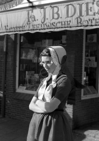 Zwart-witte foto van een jonge vrouw in een oude Walcherse dracht, ze heeft haar armen over elkaar en kijkt naar iets wat links van de fotograaf gebeurt. Op de achtergrond de winkel van Diesch, te herkennen aan de luifel waarop de tekst Fa. J.B. Diesch is te lezen, met daaronder de tekst: Echte Zeeuwsche Boter.